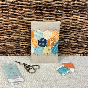 May include: A rectangular beige fabric needle case featuring a central band of colourful hexagonal patchwork in orange, blue, and yellow patterns, secured with a silver button. Includes a pack of John James quilting needles and two spools of thread.