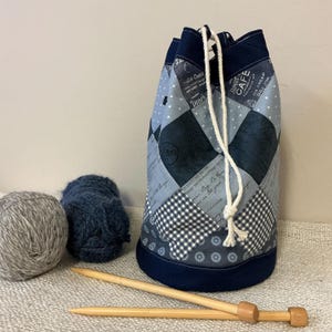 May include: A patchwork drawstring yarn bag in shades of blue, grey, and white, with a navy blue base and trim. The bag features a variety of patterns, including diamonds, polka dots, and gingham. Two balls of yarn and wooden knitting needles are next to the bag.