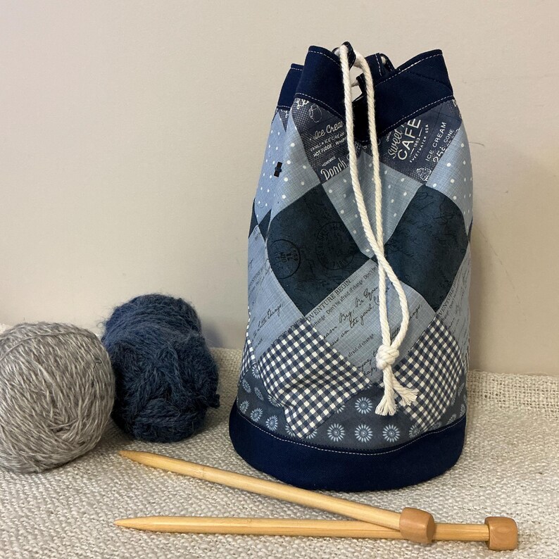 May include: A patchwork drawstring yarn bag in shades of blue, grey, and white, with a navy blue base and trim. The bag features a variety of patterns, including diamonds, polka dots, and gingham. Two balls of yarn and wooden knitting needles are next to the bag.