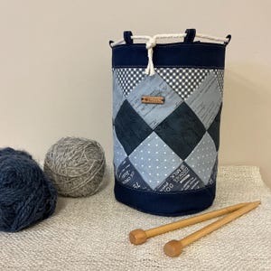 May include: A navy blue and patchwork yarn bag with a drawstring closure. The bag features a variety of gray and navy blue patterned fabrics. Two balls of yarn and wooden knitting needles are next to the bag.