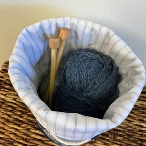 May include: A fabric knitting project bag with a blue and white striped lining. Inside the bag are two wooden knitting needles and a large ball of dark blue yarn. The bag is sitting in a woven basket.