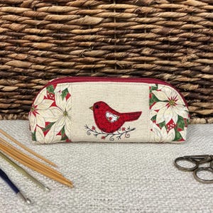 May include: A beige pencil case with a red zipper and a red bird applique. The case features panels with white poinsettias and green leaves. The bird is perched on a branch with green leaves.