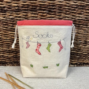 Könnte beinhalten: Ein cremefarbener Kordelzugbeutel mit rotem Musterfutter. Die Tasche hat einen gestickten Text mit der Aufschrift "Socks" über einer Wäscheleine mit bunten Socken. Grünes Gras und ein kleiner Vogel sind ebenfalls auf die Tasche gestickt.