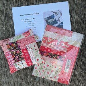 May include: A Boro stitched pin cushion featuring a patchwork of red, pink, and floral fabric scraps with white hand-stitched details, alongside a kit package containing fabric strips and an instruction sheet titled Boro Stitched Pin Cushion.