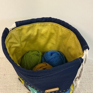 May include: A navy blue yarn bag with a yellow quilted lining. The bag holds green, blue, and brown balls of yarn. It features a white rope drawstring and a small wooden tag.
