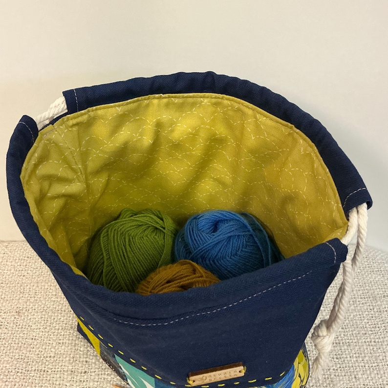 May include: A navy blue yarn bag with a yellow quilted lining. The bag holds green, blue, and brown balls of yarn. It features a white rope drawstring and a small wooden tag.