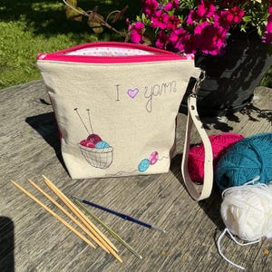 May include: A beige canvas zipper pouch with a pink interior lining and a wrist strap. The front features embroidered text that reads I heart yarn, a basket filled with yarn balls, and two knitting needles. Three wooden needles and a crochet hook.