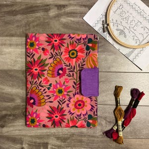 May include: A floral fabric notebook cover with a purple closure. The cover features a pink background with colorful flowers in shades of pink, orange, and yellow. Embroidery floss and a wooden hoop are also visible.