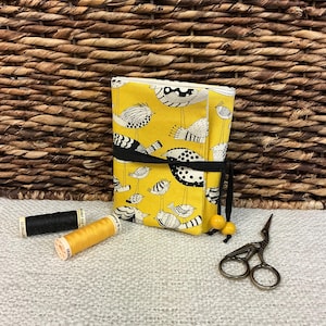 Peut inclure: Une boîte à aiguilles en tissu jaune avec une fermeture en ruban noir et un motif d'oiseaux noir et blanc. La boîte est posée sur une surface en osier tressé avec deux bobines de fil et une petite paire de ciseaux.