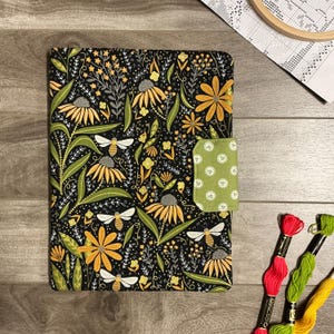 May include: A rectangular fabric cover featuring a black background with a repeating pattern of yellow flowers, green leaves, and white bees. The cover includes a light green tab closure with a white circular pattern and is made of fabric.