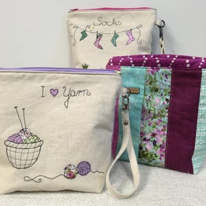 May include: Three zippered pouches with fabric handles. The pouches are made of natural linen fabric and feature embroidered designs. One pouch has the text "I love yarn" with a heart and a basket of yarn. Another pouch has the text "Socks" with a line of socks hanging on a clothesline. The third pouch has a patchwork design with pink, green, and blue fabric.