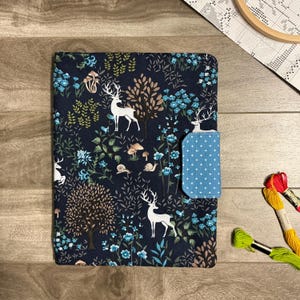 May include: A rectangular fabric book cover featuring a dark navy blue background with a repeating pattern of white deer, brown trees, blue flowers, mushrooms, and snails. A light blue tab with a white star pattern secures the cover.