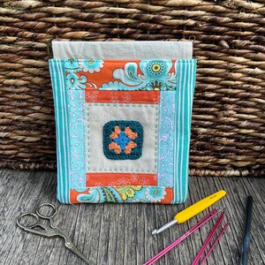 May include: A patchwork fabric organizer with a crocheted flower detail. The design incorporates orange and teal floral patterns, teal striped borders, and a central crocheted square. Crochet hooks and scissors are also visible.