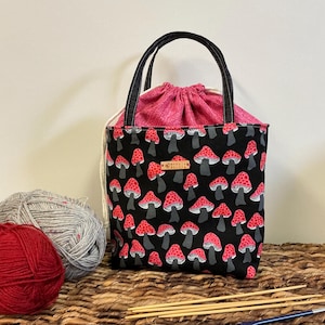 May include: A black tote bag with a red mushroom pattern and black handles. The bag has a pink drawstring closure and a small wooden tag. Two balls of yarn and knitting needles are visible.