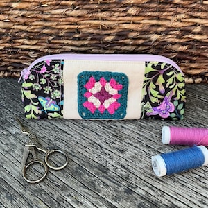 May include: A handmade pencil case with a crocheted square in the center. The case features a floral pattern on the sides and a lavender zipper. A pair of scissors and spools of thread are next to the case.