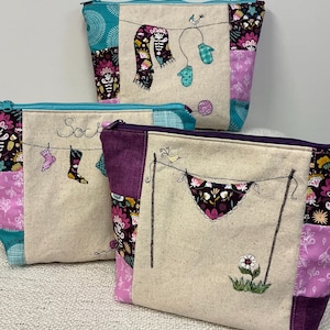 May include: Three zippered pouches made of patchwork fabric with different embroidered designs. The top pouch has a teal, pink, and purple patchwork design with an embroidered scarf and mittens. The middle pouch has a teal, pink, and purple patchwork design with the word "Socks" embroidered on it. The bottom pouch has a purple, pink, and teal patchwork design with an embroidered pair of underwear hanging on a clothesline.
