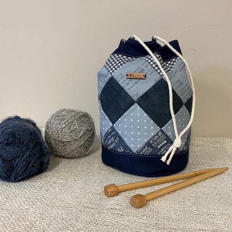 May include: A patchwork drawstring yarn bag in shades of blue and grey, with a white drawstring. Two balls of yarn and two wooden knitting needles are next to the bag. The bag has a small wooden tag.