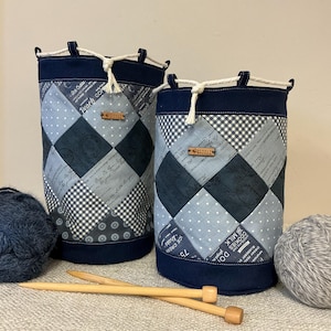 Op de afbeelding: Twee cilindrische stoffen breiprojecttassen met patchworkontwerpen in tinten blauw, grijs en wit. Elke tas heeft een marineblauwe boven- en onderkant, met een trekkoordsluiting. Breinaalden en bollen wol zijn zichtbaar.