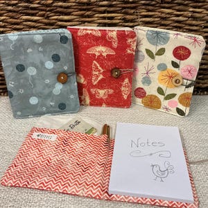 Puede incluir: Tres diarios cubiertos de tela con cierres de botón. Uno es gris con diseños azules y blancos, otro es rojo con patrones de mariposas y el tercero es crema con diseños florales. Un diario abierto con un patrón de chevrón rojo y blanco está en primer plano, con un bloc de notas que dice "Notes".