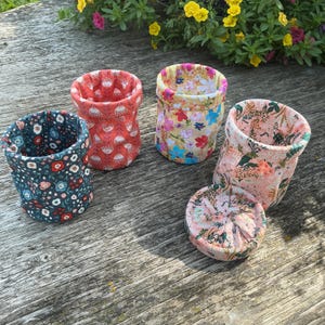 May include: Four fabric baskets with floral patterns in various colours including dark blue, coral, yellow, and light pink. Three baskets are upright and cylindrical, while one light pink basket is folded flat into a circular shape.