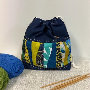 May include: A navy blue drawstring bag with a patchwork design in shades of blue, green, and yellow. The bag has a white drawstring closure, suitable for storing knitting or crochet supplies. Wooden knitting needles and yarn are visible.