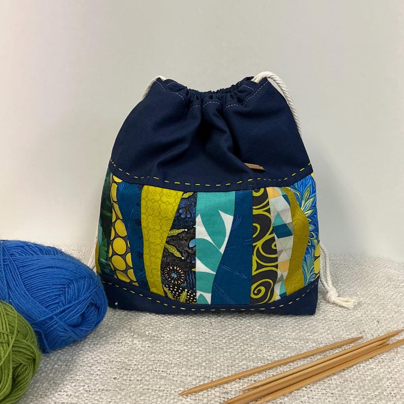 May include: A navy blue drawstring bag with a patchwork design in shades of blue, green, and yellow. The bag has a white drawstring closure, suitable for storing knitting or crochet supplies. Wooden knitting needles and yarn are visible.