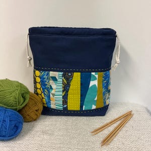 Pode incluir: Uma bolsa de fio azul marinho com cordão e um design de patchwork colorido. O patchwork apresenta vários padrões em tons de azul, verde e amarelo. A bolsa é mostrada com novelos de lã em verde, castanho e azul, e um par de agulhas de tricô de madeira.