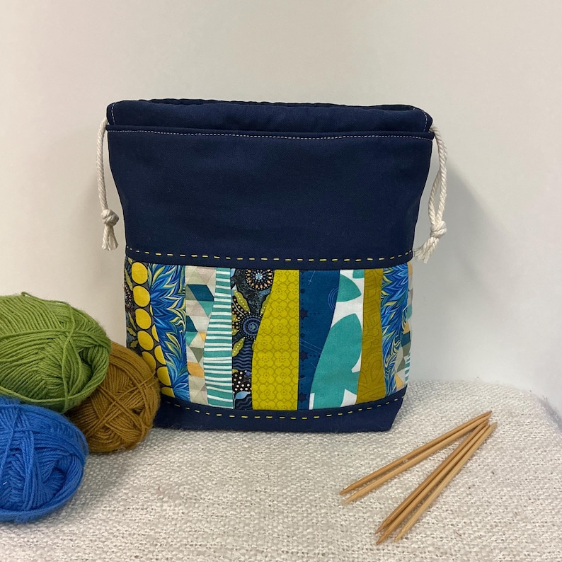 May include: A navy blue drawstring yarn bag with a colourful patchwork design. The patchwork features various patterns in shades of blue, green, and yellow. The bag is shown with balls of yarn in green, brown, and blue, and a pair of wooden knitting needles.