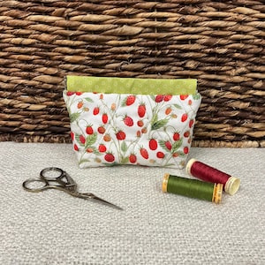 Peut inclure: Une pochette en tissu matelassé à motif de fraises rouges et de feuilles vertes sur fond blanc, surmontée d'une bande de tissu vert lime. À côté se trouvent une paire de ciseaux en métal et deux bobines de fil rouge et vert olive.
