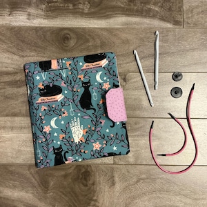 May include: A teal blue fabric project bag with a pink flap and black cats, stars, and moons. The bag has the text "The Alchemist's Practical Potions" on it. The bag is next to two white crochet hooks, two black buttons, and a pink cord.
