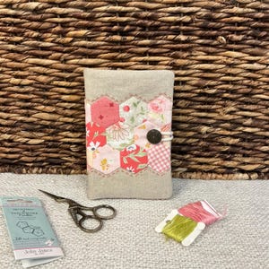 May include: A rectangular fabric needle case with a beige linen cover featuring a central band of multicoloured floral and gingham hexagon patches. The case is secured with a white elastic loop and a dark metal button, accompanied by sewing needles and thread.