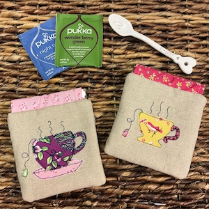 May include: Two embroidered tea cozies with teacup designs, alongside tea bags and a white spoon. One cozy features a purple teacup, the other a yellow teacup. The cozies are beige with pink floral linings. Tea bags include 'Night Time' and 'Wonder Berry Green'.