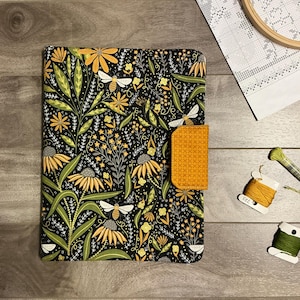 May include: A rectangular fabric cover featuring a black background with a repeating pattern of yellow flowers, green leaves, and white bees. The cover includes a solid mustard yellow tab closure with a cross-stitch texture on the right side.