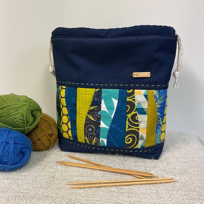 May include: A navy blue drawstring yarn bag with a patchwork design in shades of teal, yellow, and blue. The bag has a wooden tag and is next to balls of yarn and knitting needles.