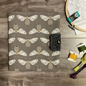 May include: A rectangular fabric cover featuring a repeating pattern of bees with yellow and black striped bodies and white wings on a grey background. A black tab with a small gold bee embroidery secures the side. Embroidery floss and a hoop are nearby.