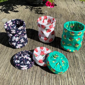 May include: Three fabric storage baskets and three matching circular fabric lids. One basket is navy blue with white flowers, one is white with red strawberries, and one is green with small red flowers. Each basket has a folded top edge.