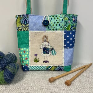 May include: A patchwork tote bag featuring various blue and green fabric patterns, including polka dots and geometric shapes. The centre panel displays an embroidered illustration of a person knitting, with a small flower nearby. A dark blue button closure secures the top, and two fabric handles are attached. A ball of blue and green yarn and two wooden knitting needles are positioned in front.