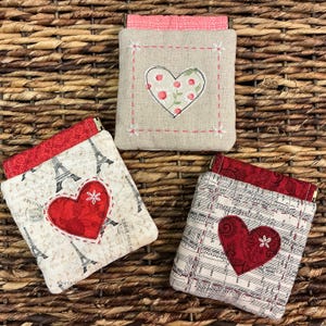 May include: Three small, square fabric pouches with red and white heart appliqués. One pouch features a floral heart, another has an Eiffel Tower design, and the third displays musical notes. Each pouch has a contrasting red top and a zip.