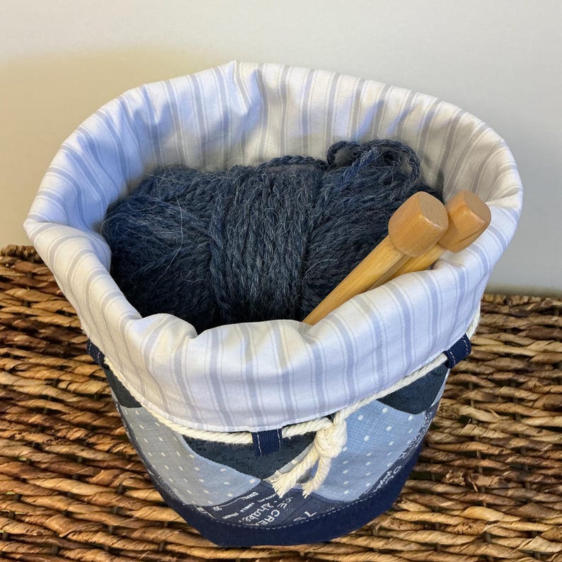 May include: A fabric yarn basket with a blue and white striped lining and a patchwork exterior. The basket holds a ball of dark blue yarn and two wooden knitting needles. A white rope detail accents the top edge.