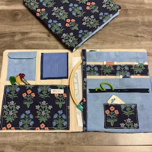 May include: A blue and floral patterned sewing organiser with multiple pockets, a zip, and a matching notebook. The organiser includes a wooden embroidery hoop, spools of thread, and green scissors. The floral pattern includes blue and orange flowers.