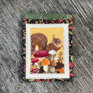 May include: A fabric notepad cover featuring a squirrel illustration surrounded by mushrooms and autumn leaves. The cover has a black background with a colorful leaf pattern and a green flap closure.