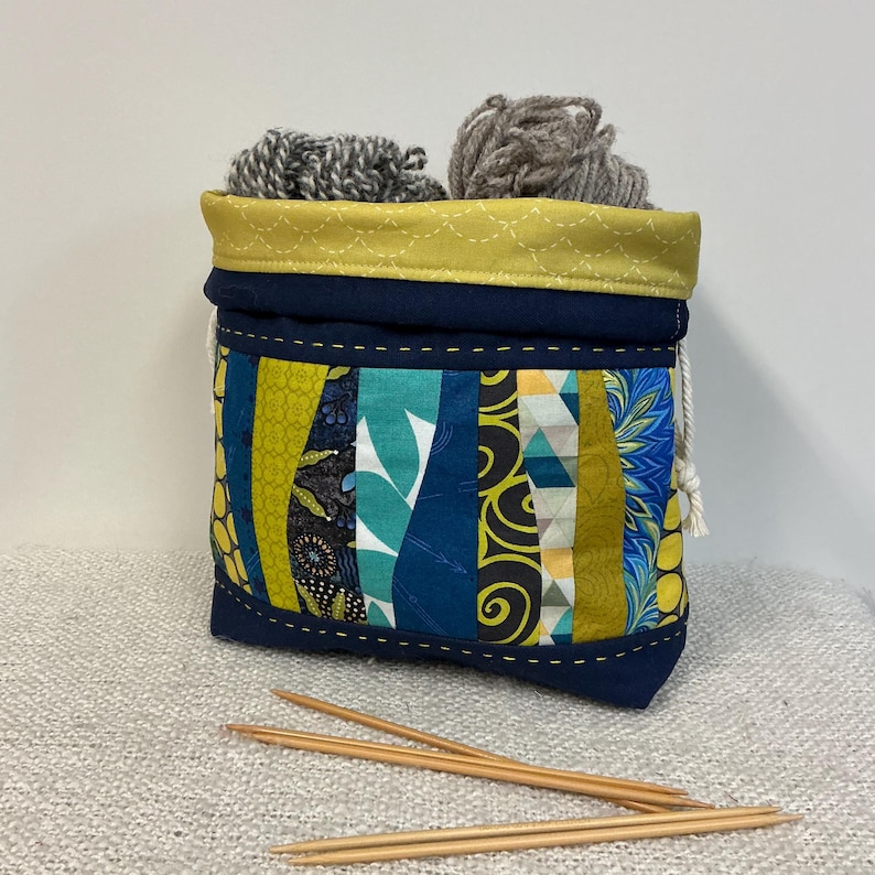 May include: A fabric yarn storage bag with a patchwork design, featuring a navy blue base and a yellow top. The bag is decorated with colourful fabric strips in blue, green, and yellow. Two balls of grey yarn are inside, with wooden knitting needles in front.