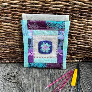 May include: A handmade, quilted crochet project bag with a square, crocheted flower design in the centre. The bag features patterned fabrics in blue, purple, and cream. Crochet hooks and scissors are visible.