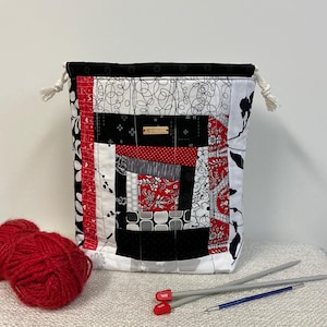 Könnte beinhalten: Ein Patchwork-Beutel mit Kordelzug in Rot, Schwarz und Weiß mit verschiedenen Mustern. Der Beutel hat eine schwarze Samtverkleidung und weiße Kordelzüge. Rote Wolle und Stricknadeln liegen neben der Tasche.