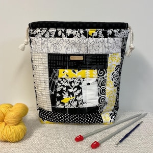 Könnte beinhalten: Eine Patchwork-Garntasche mit Kordelzugverschluss. Die Tasche hat eine Mischung aus schwarzen, weißen und gelben Stoffen mit verschiedenen Mustern, darunter Blumen-, geometrische und Lineal-Designs. Gelbes Garn und Stricknadeln liegen neben der Tasche.