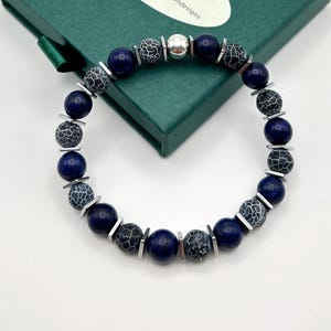 Könnte beinhalten: Ein Armband mit dunkelblauen und schwarz gemusterten Perlen, unterbrochen von silberfarbenen Metallakzenten. Das Armband ist kreisförmig und liegt auf einer grünen Schachtel. Die Perlen haben einen Durchmesser von etwa 1,3 cm.