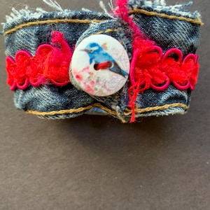 May include: A denim cuff bracelet with a button featuring a blue bird design. The bracelet is accented with bright red trim and pink thread details. The cuff is approximately 2 inches wide.