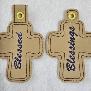 May include: Two tan cross-shaped keychains with dark brown stitching. One keychain says "Blessed" in blue script, and the other says "Blessings". Each has a yellow grommet.