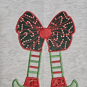May include: Embroidered Christmas elf design with green and red striped legs and green elf shoes. The elf is wearing a red and white candy cane bow.
