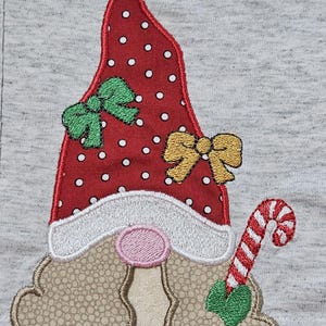 May include: Embroidered gnome with a red and white polka dot hat, green and yellow bows, a white beard, and a pink nose. The gnome is holding a candy cane in its right hand.
